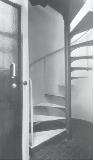 Schwarz-weiß-Foto einer engen Spindeltreppe hinter geöffneter Tür, mit Geländer und dunklem Fliesenboden, Corbusier